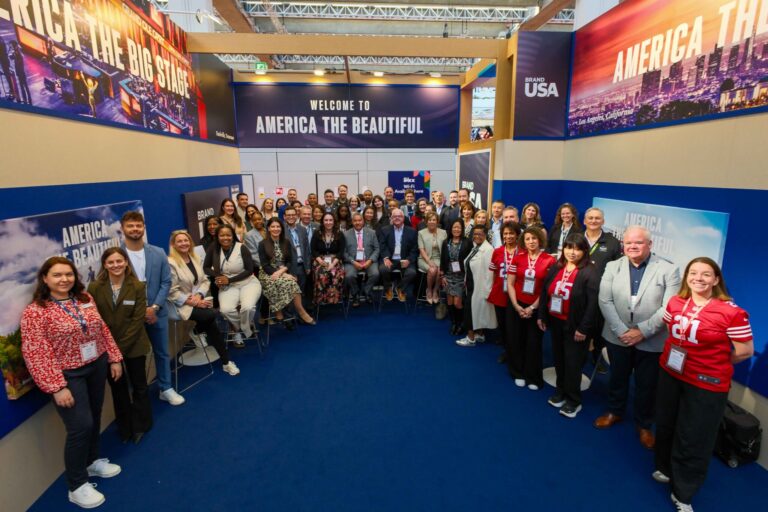 A large group of people pose and smile in a conference booth decorated with American imagery and the words “Welcome to America the Beautiful.” Some wear red sports jerseys, others are in business attire.
