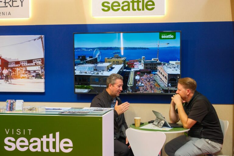 Two men sit at a booth labeled "Visit Seattle," talking with a laptop and coffee cup between them. A large screen behind shows an aerial view of Seattle with the waterfront and the Ferris wheel.