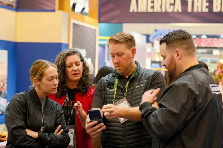 Four adults stand close together at an event, looking at a phone one man is holding. They appear focused and engaged in conversation. Booths and signs are visible in the busy background.