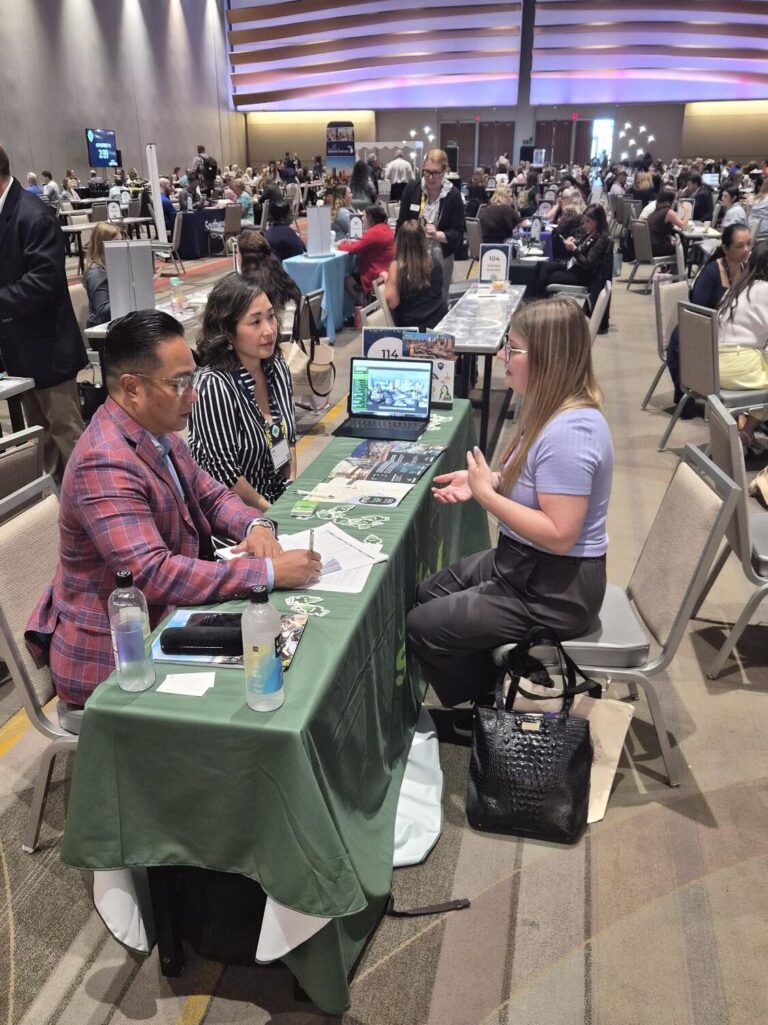 A woman talks to two people seated behind a table at a busy conference or networking event. Papers, a laptop, and a water bottle are on the table. Many others are visible meeting in the background.