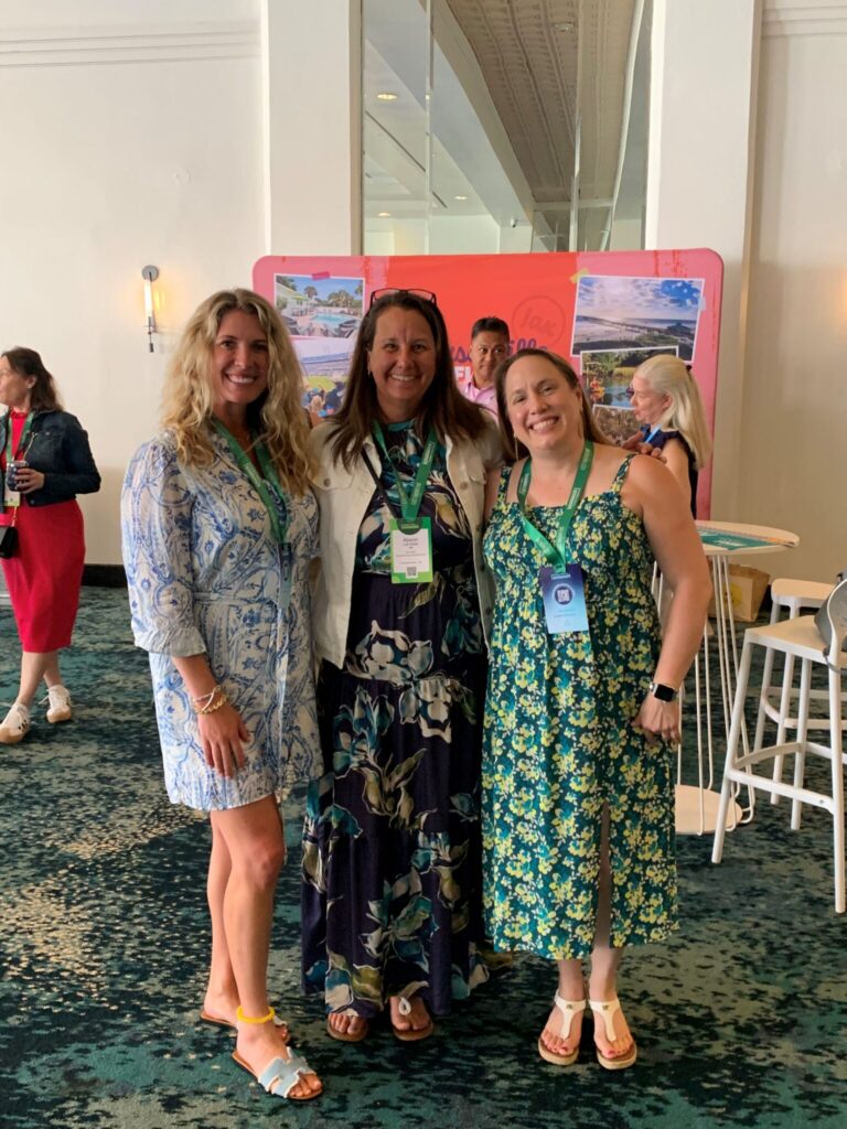 Three women in summer dresses stand smiling indoors at an event, wearing name badges. Other people are in the background near tables and a colorful backdrop with images and text.