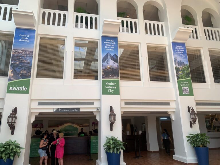 Hotel lobby with registration desk, two women talking, and "Visit Seattle" banners promoting fresh air, Mother Nature’s City, and outdoor exploration. Large windows and plants decorate the bright, open space.