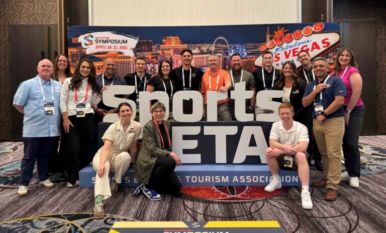 A group of people pose and smile together in front of a large “Sports ETA” sign and a Las Vegas backdrop at a symposium event.