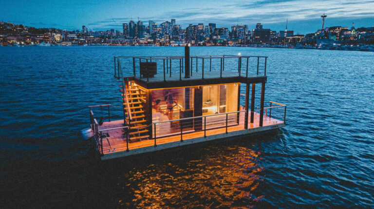 A modern floating house with large windows and rooftop deck sits on calm water at dusk, with a city skyline glowing in the background.
