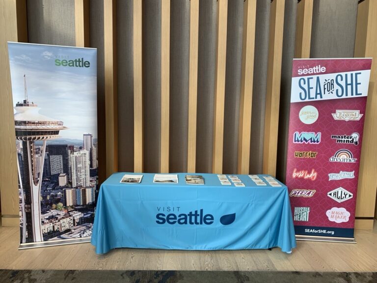 A Visit Seattle booth with a blue tablecloth, brochures on the table, and two standing banners—one with the Space Needle and cityscape, the other promoting SEA for SHE with various logos.