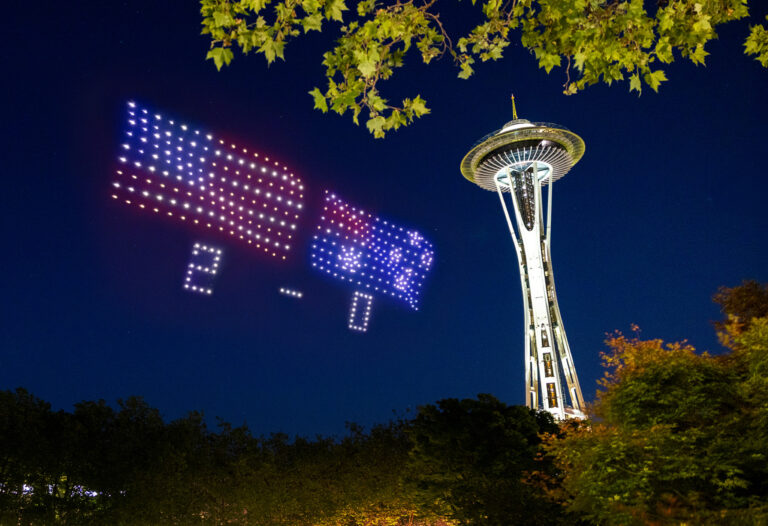 Drones form images of the US and Australian flags, and the numbers “2-0,” next to the Space Needle at night, with green leaves and trees in the foreground.