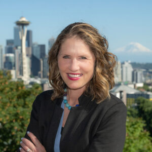 A woman with wavy brown hair smiles in front of the Seattle skyline, with the Space Needle and Mount Rainier visible in the background on a clear, sunny day. She is wearing a black jacket and a blue necklace.