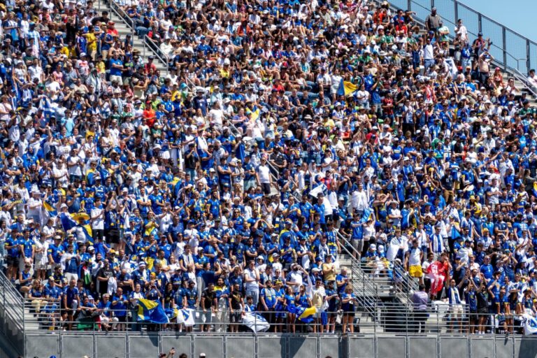 A large crowd of fans dressed mostly in blue and white sit and stand in stadium bleachers, cheering and waving flags under a clear, sunny sky.