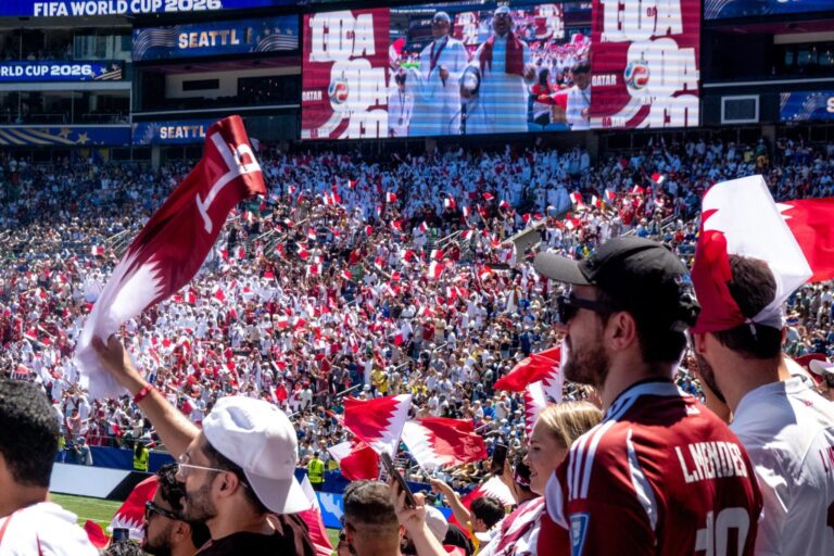 A stadium crowd waves Qatar flags, many dressed in white. A large screen displays people with flags. "FIFA World Cup 2026" and "SEATTLE" banners are visible above the stands.