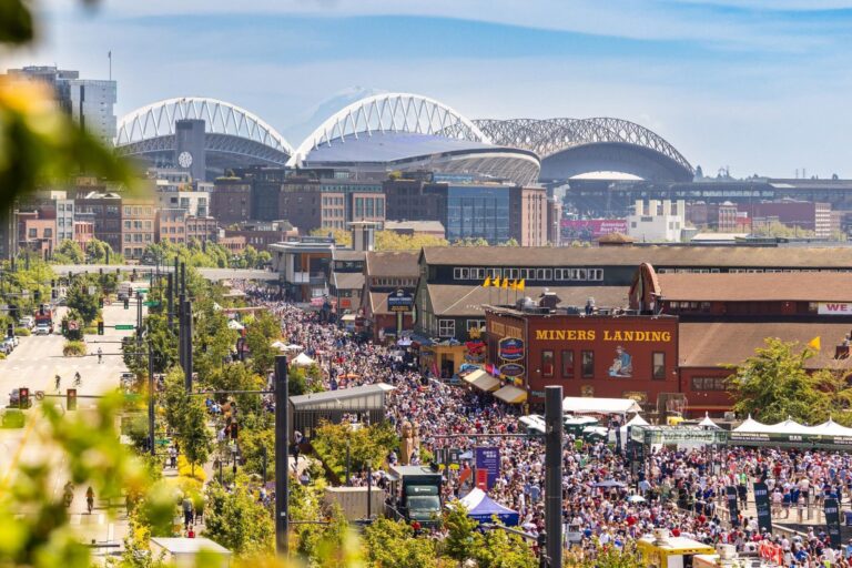A large crowd gathers at Miners Landing waterfront in Seattle on a sunny day, with stadiums in the background and city buildings visible.