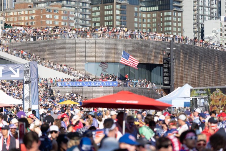A large crowd gathers outdoors near an urban building, with an American flag flying above. People are wearing hats and sunglasses, and city buildings are visible in the background.