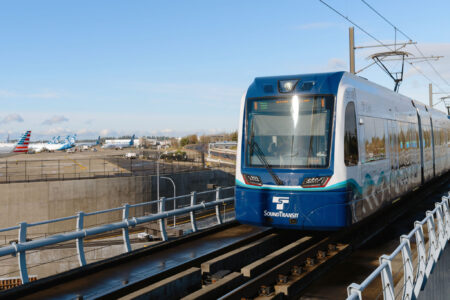 A Sound Transit light rail train approaches on elevated tracks with an airport and airplanes visible in the background under a clear sky.