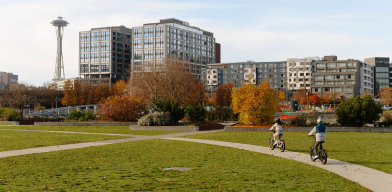 Two people ride bikes on a path through a grassy park with autumn trees, modern buildings, and the Space Needle visible in the background.