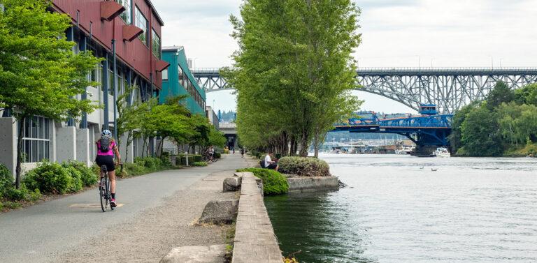 A cyclist rides on a riverside path lined with trees and buildings, with people sitting nearby. A large steel bridge spans the river in the background under a cloudy sky.