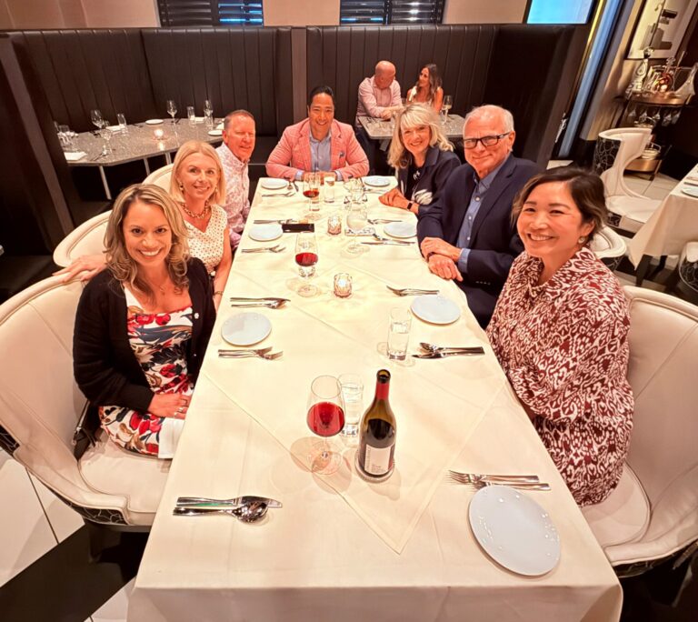 Seven people sit around a white tablecloth-covered table at a restaurant, smiling at the camera. There are drinks, plates, and utensils on the table. The background shows other diners and a stylish, modern decor.