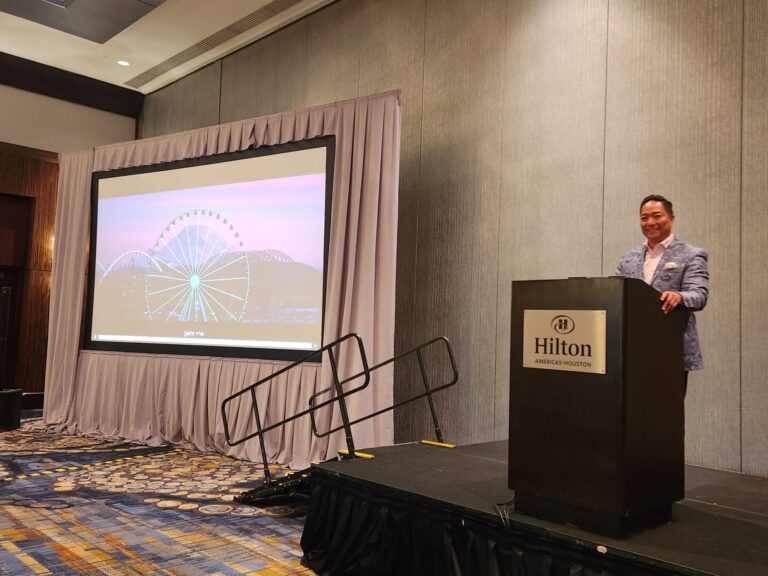 A man stands at a podium labeled "Hilton Americas-Houston" beside a screen displaying an image of a ferris wheel at sunset in a conference room.