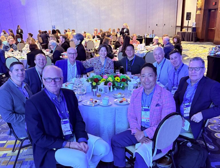 A group of people in business attire sit around a round table with flowers and teacups at a conference event, while others are seated at tables in the background.