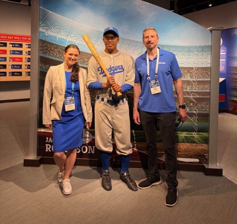 Three people pose indoors; two stand on either side of a life-sized statue of a baseball player in a Dodgers uniform, holding a bat, with a stadium backdrop behind them.