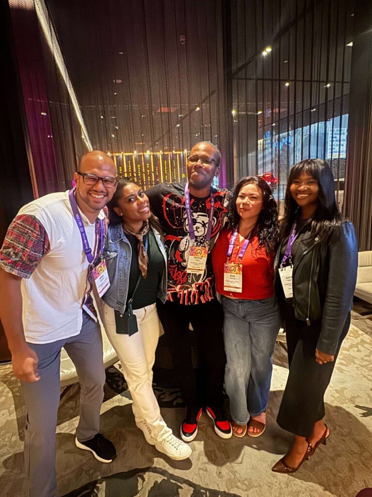 Five people stand close together indoors, smiling at the camera. They wear conference badges and casual to business-casual attire. Curtains and warm lighting appear in the stylish background.