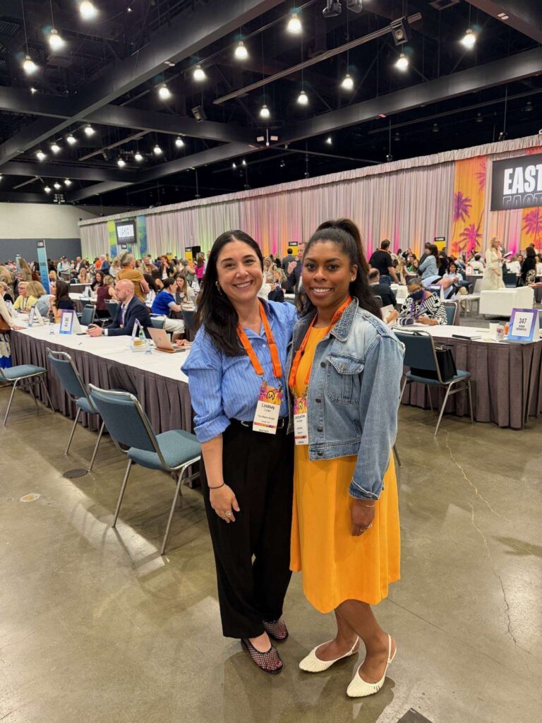 Two women smile and pose together at a conference. They wear name badges, and many people are seated at tables in the background in a large, well-lit event space with colorful signage.