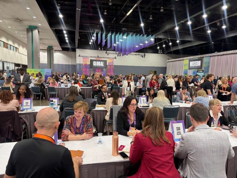 Large conference hall with many people seated at tables having discussions. Signage for “GLOBAL” and “WESTERN” hangs in the background, and the atmosphere is lively and professional.