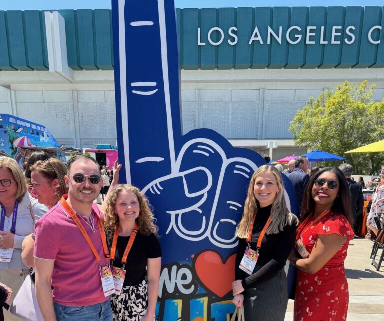 A group of smiling people pose outdoors by a large foam finger at an event in front of the Los Angeles Convention Center.