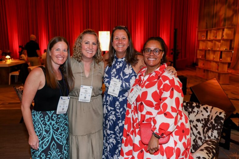 Four women smiling and posing together at an indoor event with red curtains in the background, all wearing name badges and colorful dresses.