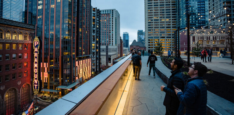 People stand on a rooftop terrace overlooking a cityscape with tall buildings, including the illuminated Paramount sign. The sky is overcast and string lights hang above the terrace.