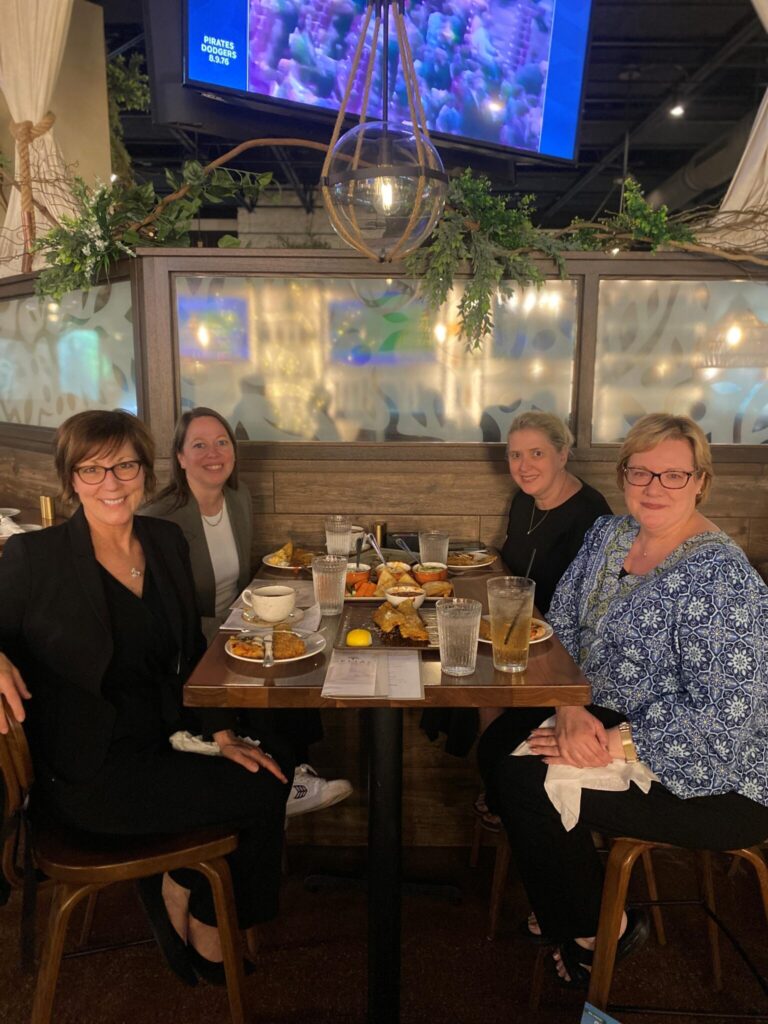 Four women sit around a restaurant table with plates of food and drinks, smiling at the camera. The setting is warmly lit with greenery and decorative lighting above them.