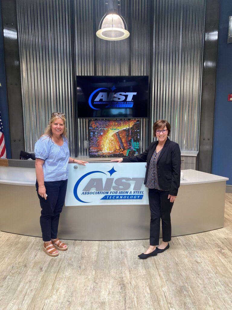 Two women stand and smile in front of a reception desk with an “Association for Iron & Steel Technology” sign, in a modern lobby with a TV displaying industrial imagery.