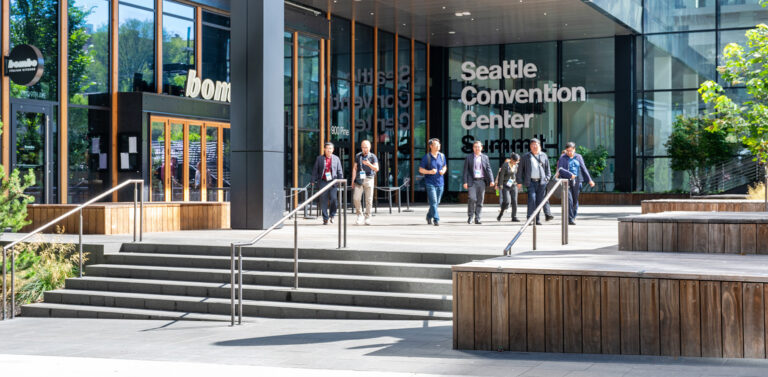 People walk down steps outside the Seattle Convention Center on a sunny day. Large glass windows and modern architecture are visible, with greenery and wooden benches in the foreground.