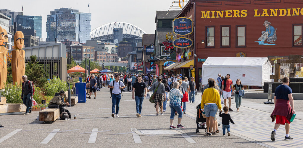 People walk along a busy waterfront with shops, restaurants, and signs, including “MINERS LANDING.” The scene includes greenery, city buildings, and a distant stadium.