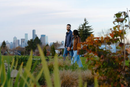 A couple walks hand in hand through a park with autumn foliage, with a city skyline and tall buildings visible in the background under a partly cloudy sky.