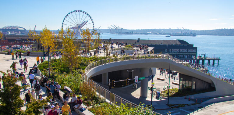 People stroll along a waterfront park with lush landscaping. There is a large Ferris wheel, piers, and ships in the harbor on a sunny day. A modern elevated walkway curves above the scene.