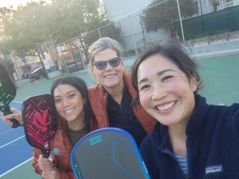 Three women smiling and posing on an outdoor tennis or pickleball court, each holding a paddle. The court is surrounded by a fence, with trees and buildings visible in the background.