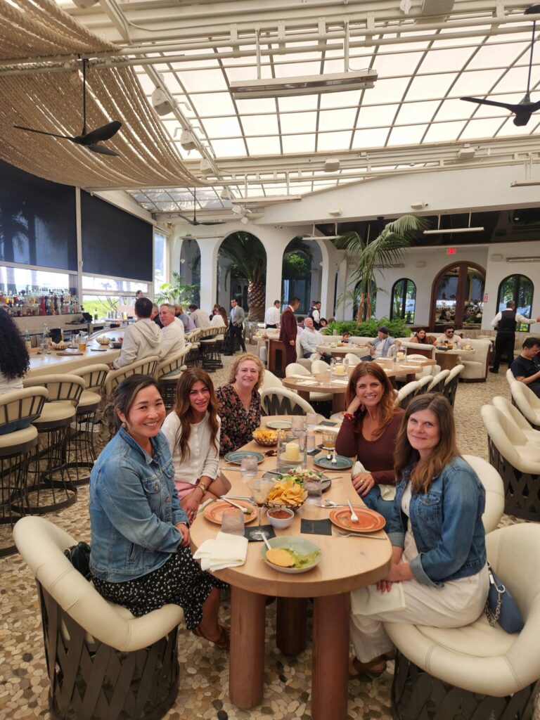 Six women sit around a round table at a stylish restaurant, smiling at the camera. Food and drinks are on the table. The restaurant has a bright, airy design with plants and modern decor.