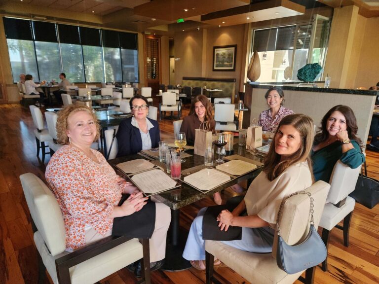 Six women sit around a restaurant table with menus, drinks, and glassware, smiling at the camera. The restaurant has wooden floors, white chairs, and large windows letting in natural light.