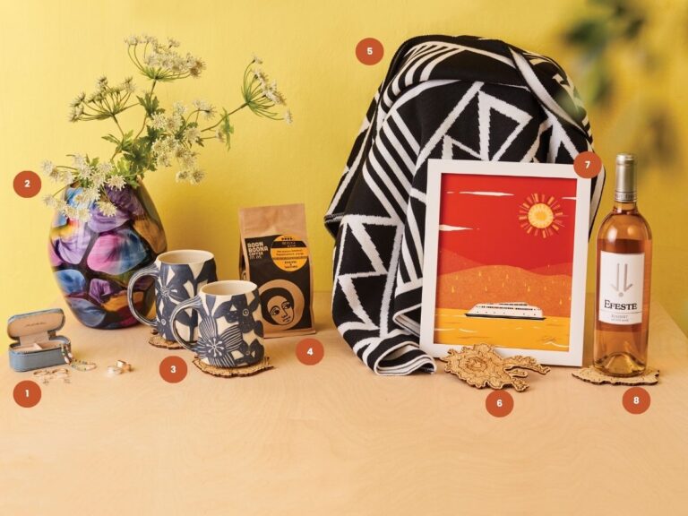 Table with colorful vase of flowers, two mugs, coffee bag, jewelry box, geometric blanket, framed art of a boat, wine bottle, and decorative coaster, all set against a yellow wall.