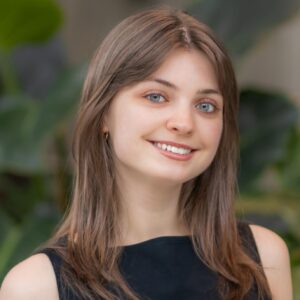 A young woman with straight brown hair and blue eyes smiles at the camera, wearing a sleeveless black top. The background is blurred with green leafy plants.