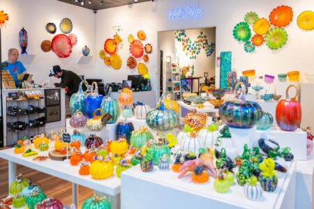 A bright glass art shop displays colorful glass pumpkins, bowls, and wall art. Two people stand behind the counter. The white walls and shelves highlight the vibrant, handcrafted glass pieces throughout the store.