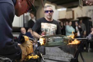 A person shapes a hot glass object with a drill while another person supervises, showcasing one of the unique things to do in Seattle. Both wear protective glasses as a group of people watches in the background.