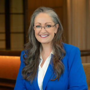 A woman with long gray hair, glasses, and a blue blazer smiles while sitting indoors in a warmly lit room.