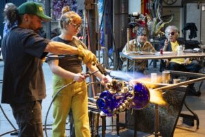 Two people use fire torches to shape a large blue glass sculpture—a mesmerizing example of the creative things to do in Seattle. In the background, three people sit at a table, watching the process and taking notes. The setting appears to be an art studio or workshop.