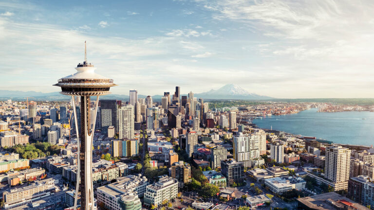 Aerial view of downtown Seattle with the Space Needle in the foreground, skyscrapers, Elliott Bay, and Mount Rainier visible in the distance under a partly cloudy sky.