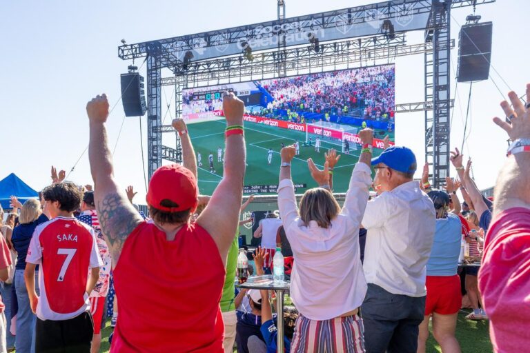 People cheer and raise their arms while watching a soccer match on a large outdoor screen at a 2026 World Cup watch party in Seattle. Some fans wear soccer jerseys, and the atmosphere is lively and festive.