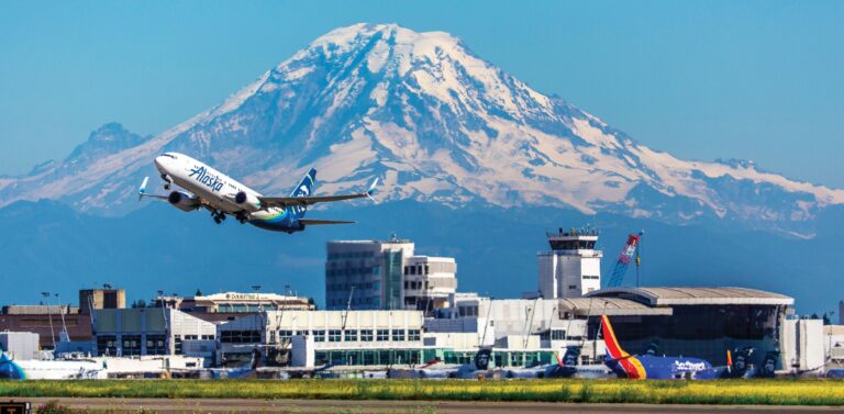 An Alaska Airlines plane takes off from an airport with snow-capped Mount Rainier in the background under a clear blue sky.