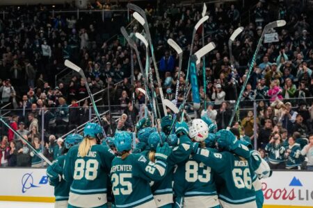 A women’s hockey team in teal uniforms huddles on the ice, raising their sticks together while a cheering crowd watches from the stands in the background.