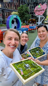 Three smiling people hold soccer-themed dioramas with moss, flags, and soccer balls. A large soccer ball display and an Elephant Super Car Wash sign are in the background at an outdoor event.
