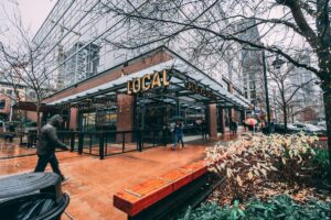 People walk in front of a modern restaurant called “LOCAL” on a rainy day. The ground is wet, some people carry umbrellas, and city buildings surround the area. Leafy plants and bare trees are visible in the foreground.
