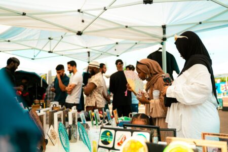 People browse art and crafts at outdoor market stalls under white tents. Two women in hijabs examine items at a table, while others chat and walk in the background. Bright, lively atmosphere.